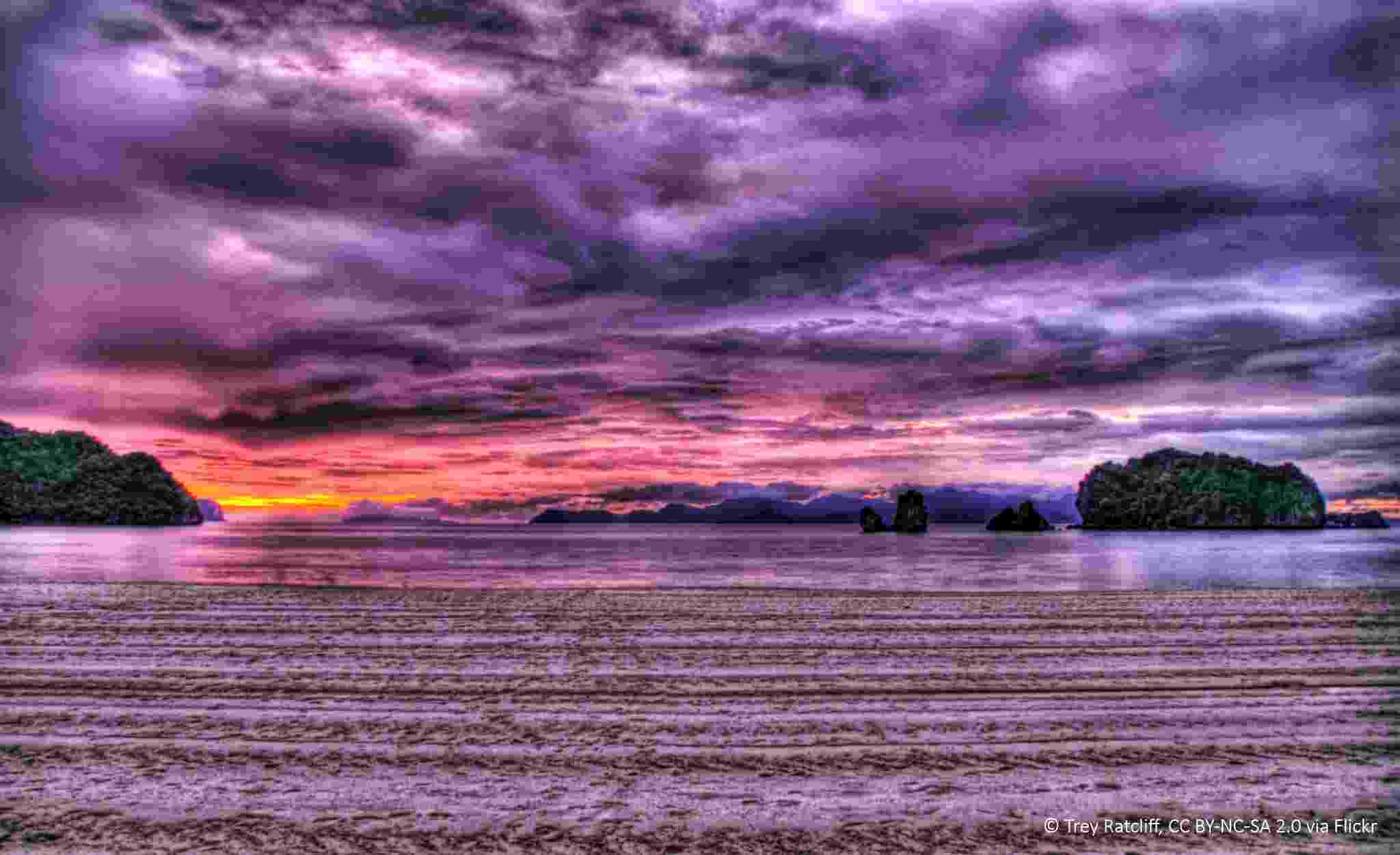 View of a purple sky and red and purple clouds across the sea, with small offshore rocks and tree-covered islands